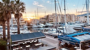 Uma marina com barcos atracados, um restaurante chamado 'L'Escalé' e edifícios ao fundo.