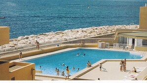 Uma piscina com pessoas nadando e tomando sol, rodeada por uma praia de pedras e vista para o mar com barcos.