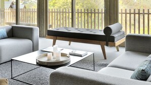 A modern interior with a sofa, a massage table, and a coffee table with candles. Large windows offer a view of a mountainous landscape.