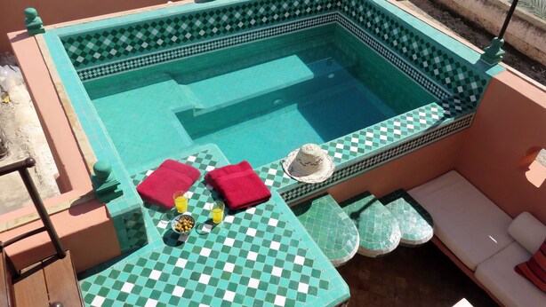 A pool area with checkered tiles, a lounge chair, a small table with a drink and snacks, and a hat on the table. - Riad "LA DOUIRIA" in exclusivity (Marrakech)