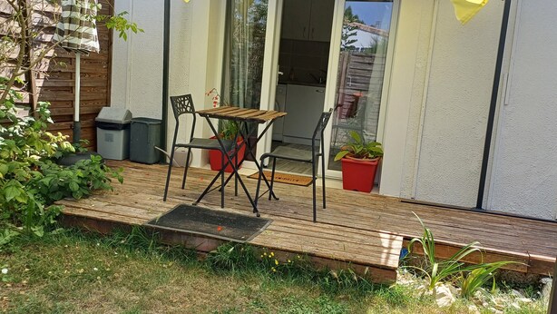 A wooden deck with a table and chairs, potted plants, and a sliding glass door leading to a kitchen. - 2* furnished studio - La Maison aux Mille Roses (Nieul-Sur-Mer)