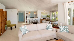 Un amplio salón con un sofá color crema, una estantería de madera y una zona de comedor con una cocina al fondo.