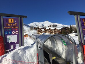 Ingang van een skilift met een bordje voor 'Tapis des Petits Lutins' en een zicht op besneeuwde gebouwen op de achtergrond.