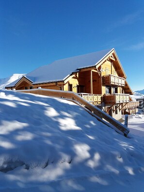 Una cabaña de madera con balcón, rodeada de terreno cubierto de nieve y montañas al fondo.