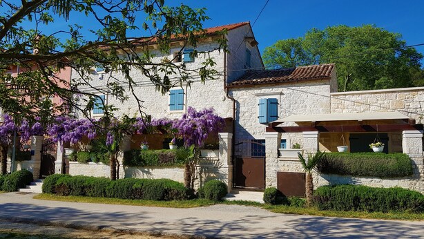 Ein Gebäude aus Stein mit traditioneller Gestaltung, mit einer überdachten Veranda und einem kleinen Garten mit violetten Blumen.