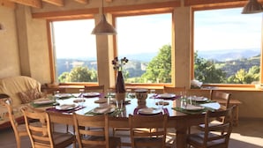 Un coin repas avec une table en bois dressée pour six personnes, entourée de chaises, et de grandes fenêtres donnant sur la campagne.