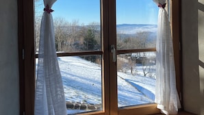 Une pièce dotée d’un plafond en bois et d’une grande fenêtre ornée de rideaux blancs, retenus par des liens, qui dévoile un paysage enneigé et des montagnes au loin.