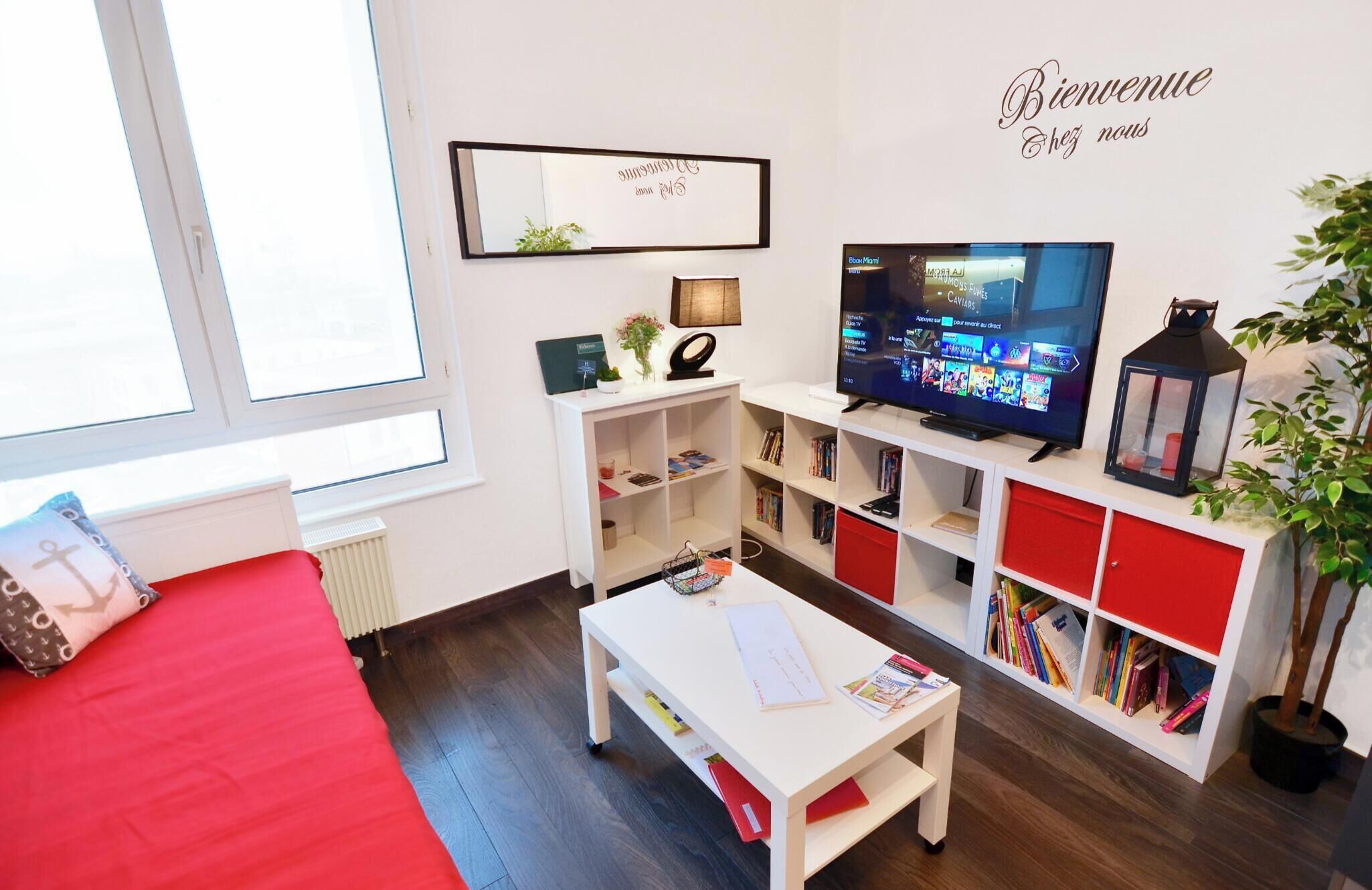 A one-bedroom apartment with a view of the port and the sea. It includes a bed, a desk, a bookshelf, and a television.