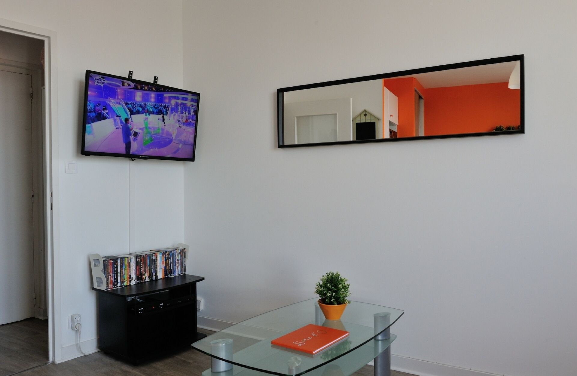 A living room with a flat-screen TV mounted on the wall, a bookshelf with DVDs, a glass coffee table with a book and a small plant, and a large mirror reflecting the room.