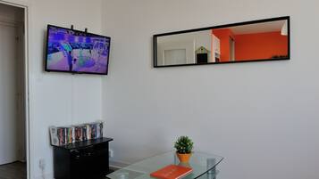 A living room with a flat-screen TV mounted on the wall, a bookshelf with DVDs, a glass coffee table with a book and a small plant, and a large mirror reflecting the room.