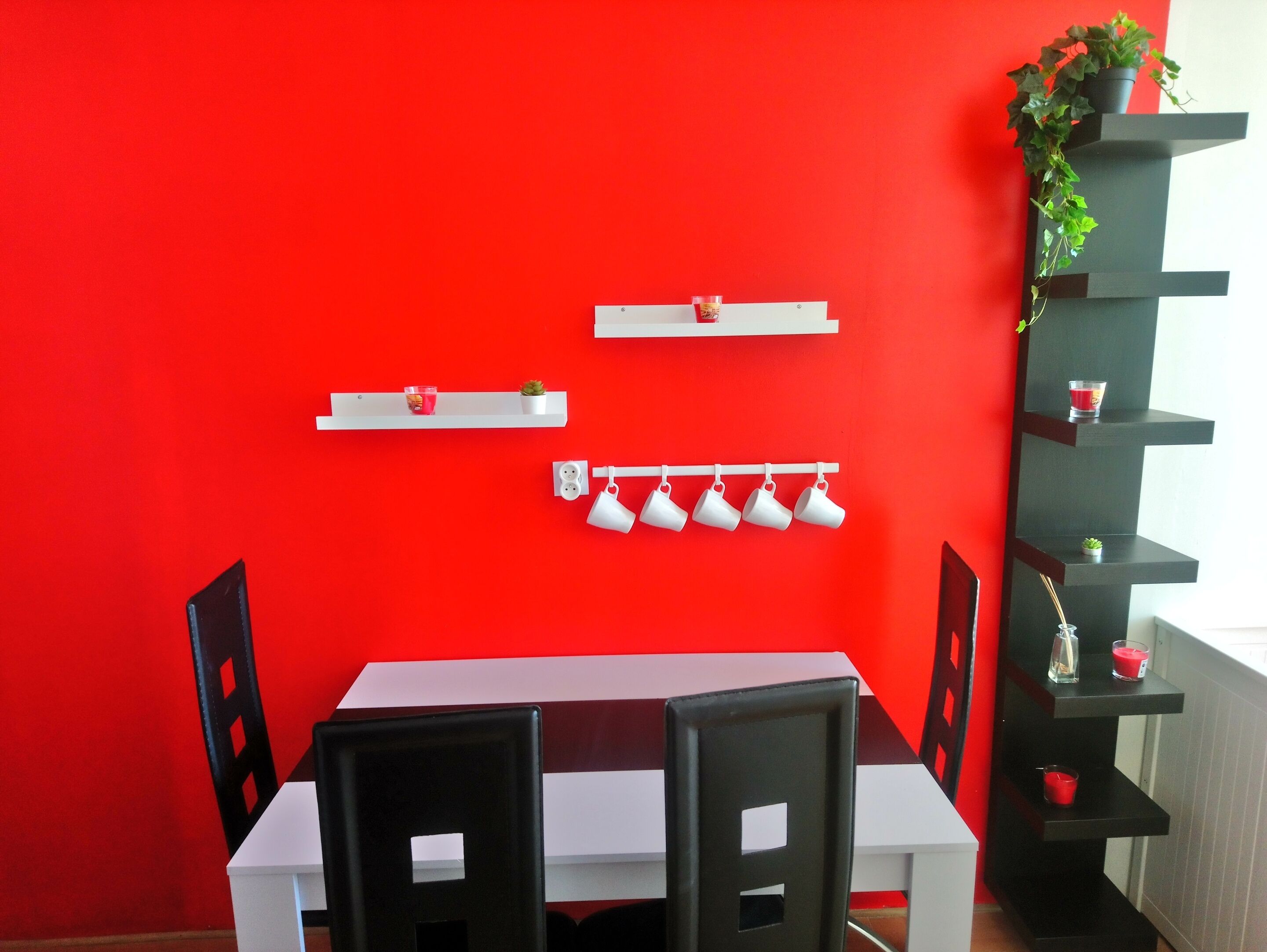 Un coin repas avec un mur rouge, une table blanche et des chaises noires. On y trouve des étagères avec des tasses et une petite plante.