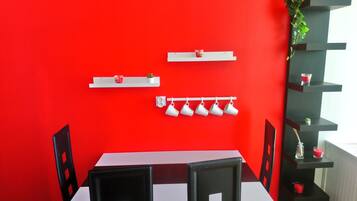 Un coin repas avec un mur rouge, une table blanche et des chaises noires. On y trouve des étagères avec des tasses et une petite plante.