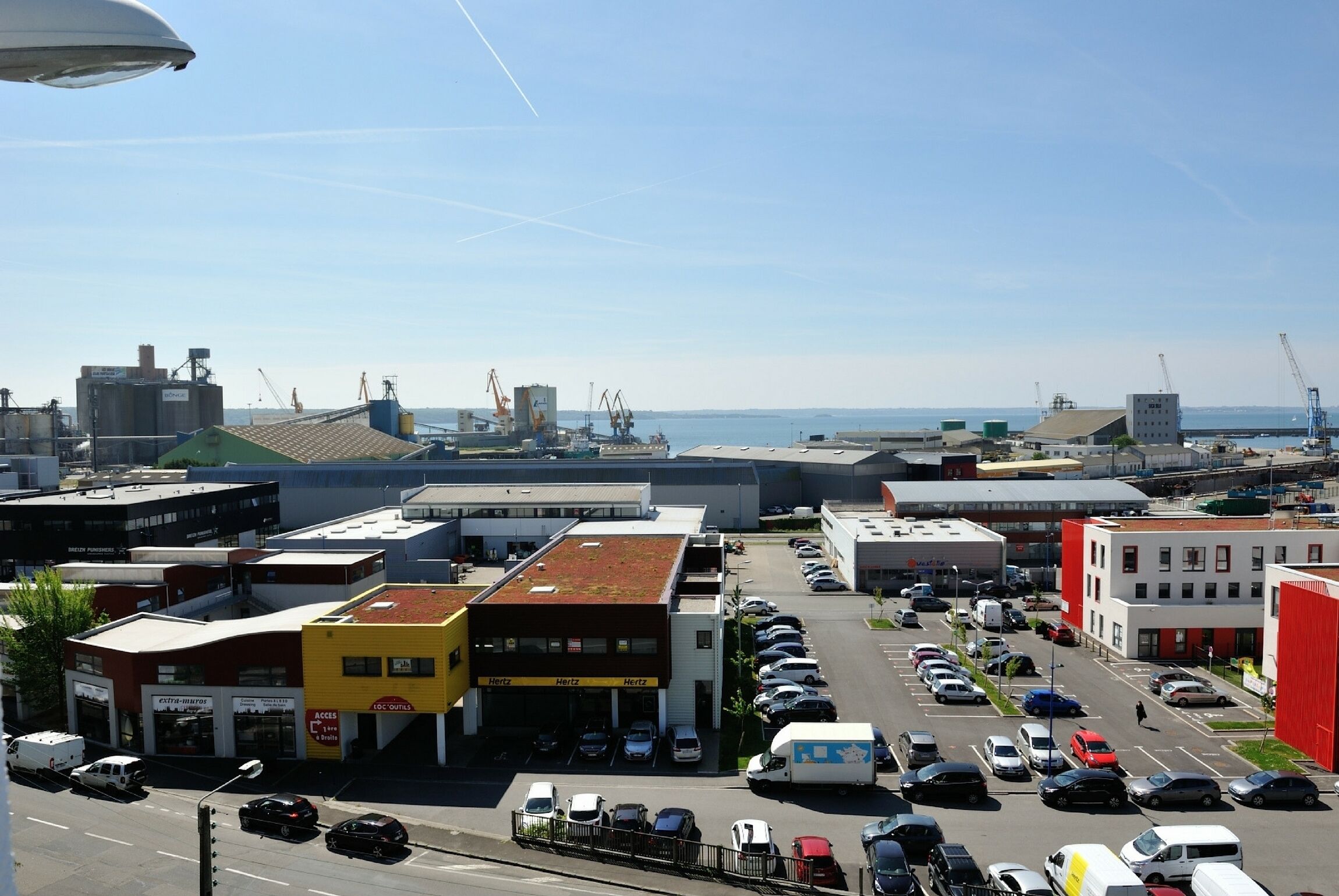 Un estacionamiento con varios autos y algunos camiones, rodeado de edificios industriales y una vista despejada del mar.