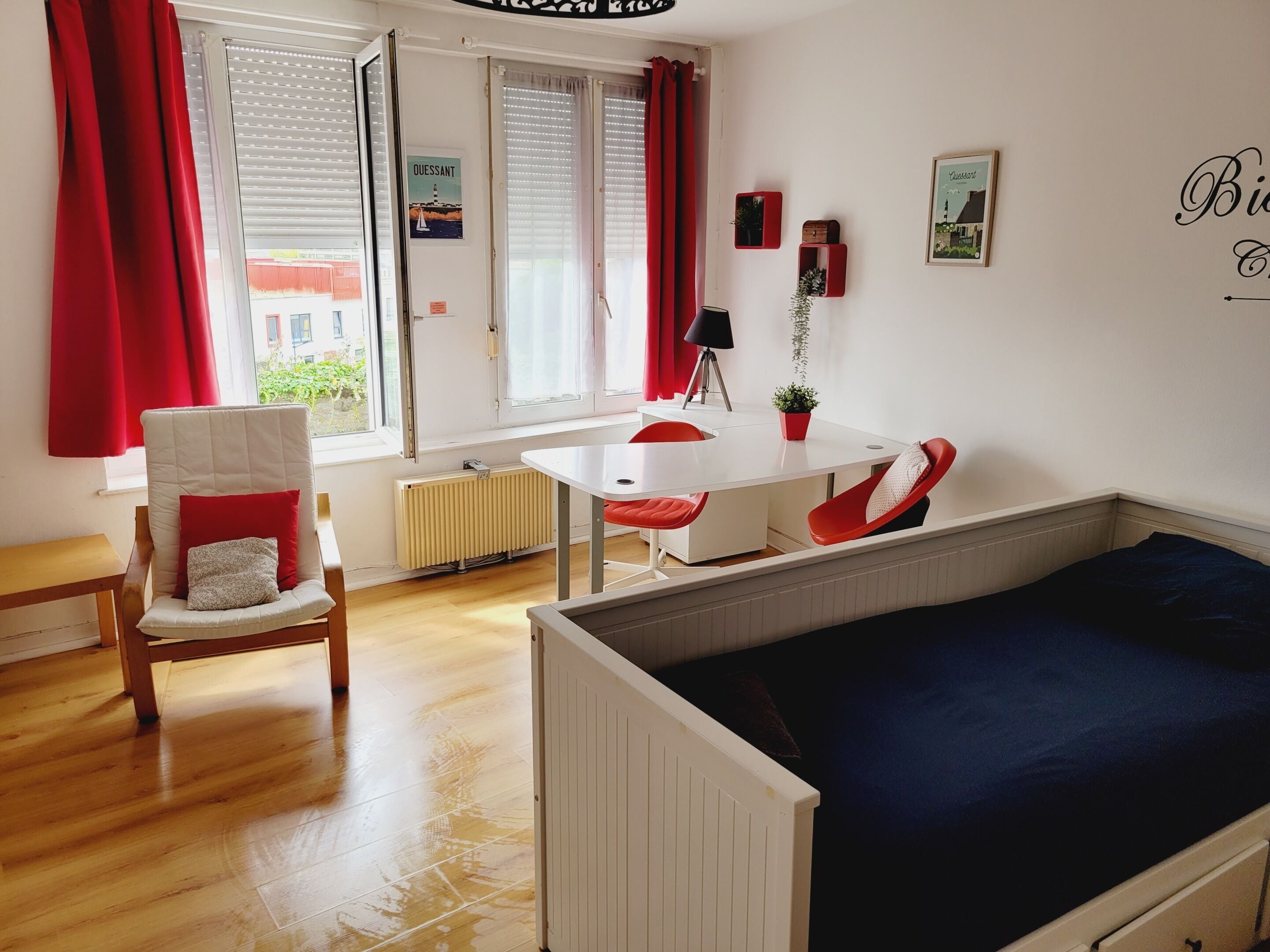Una habitación con cama, una mesa y una silla. Se ve una ventana con cortinas, un radiador y una pared decorada con cuadros.