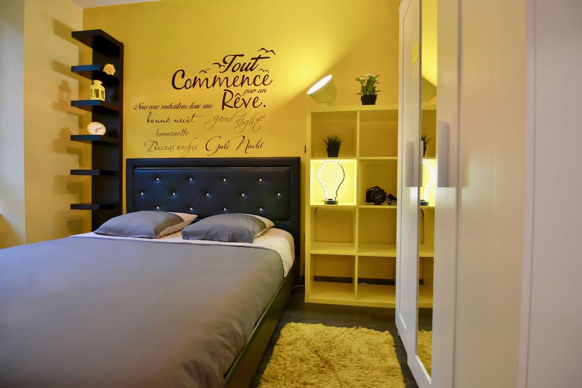 Un dormitorio con cama, una pared con una cita, una estantería con objetos y una mesita de noche con lámpara.