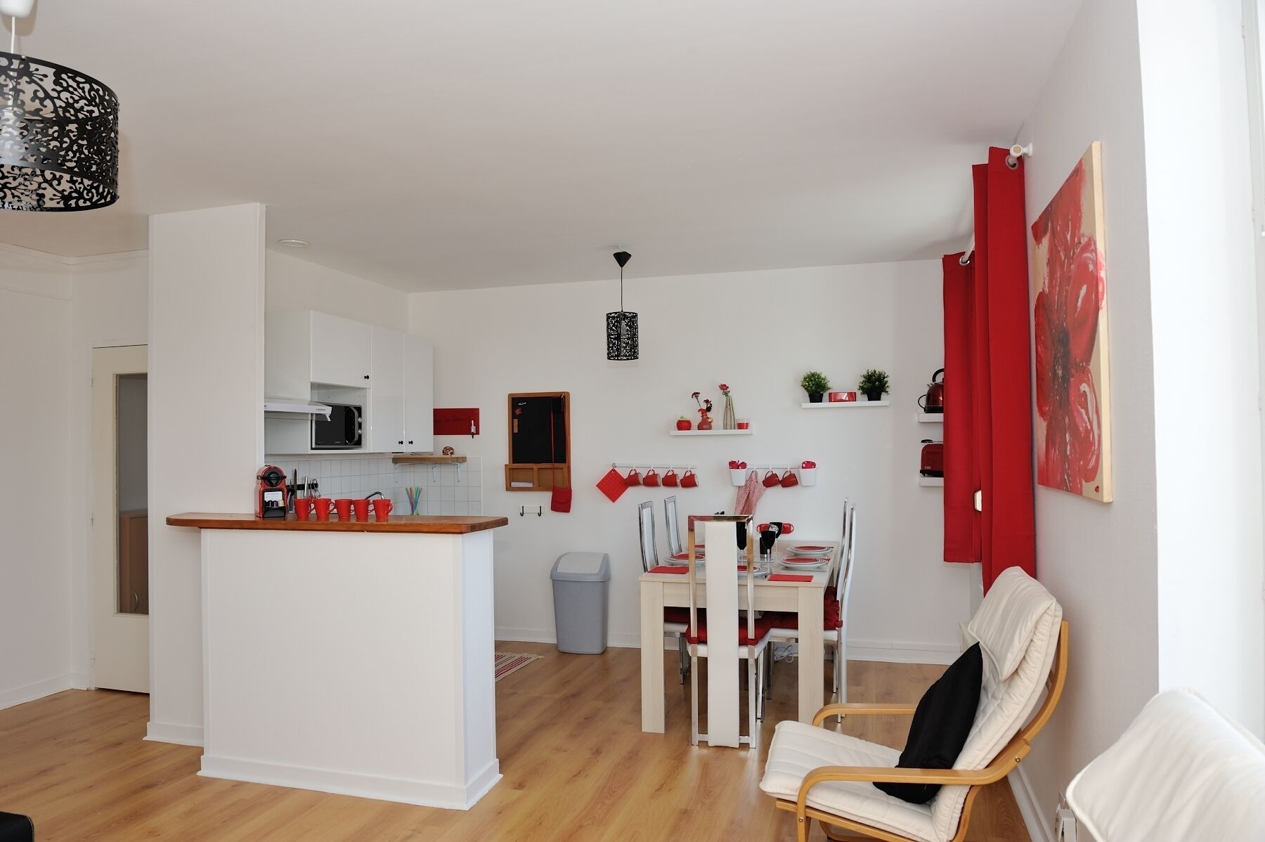 A modern kitchen with white cabinets, a central island, and red accents. There is a dining area with a white table and chairs, and a living area with a white sofa and a red curtain.