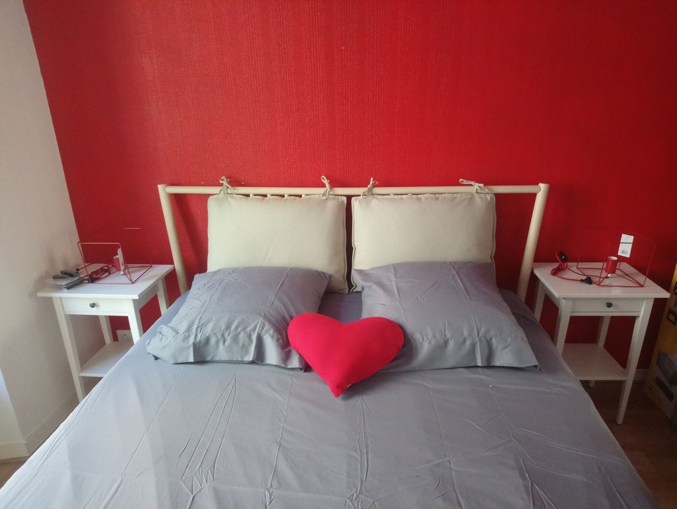 A bed with a grey bedspread and two pillows, one red and one grey, in a room with red walls and white furniture.