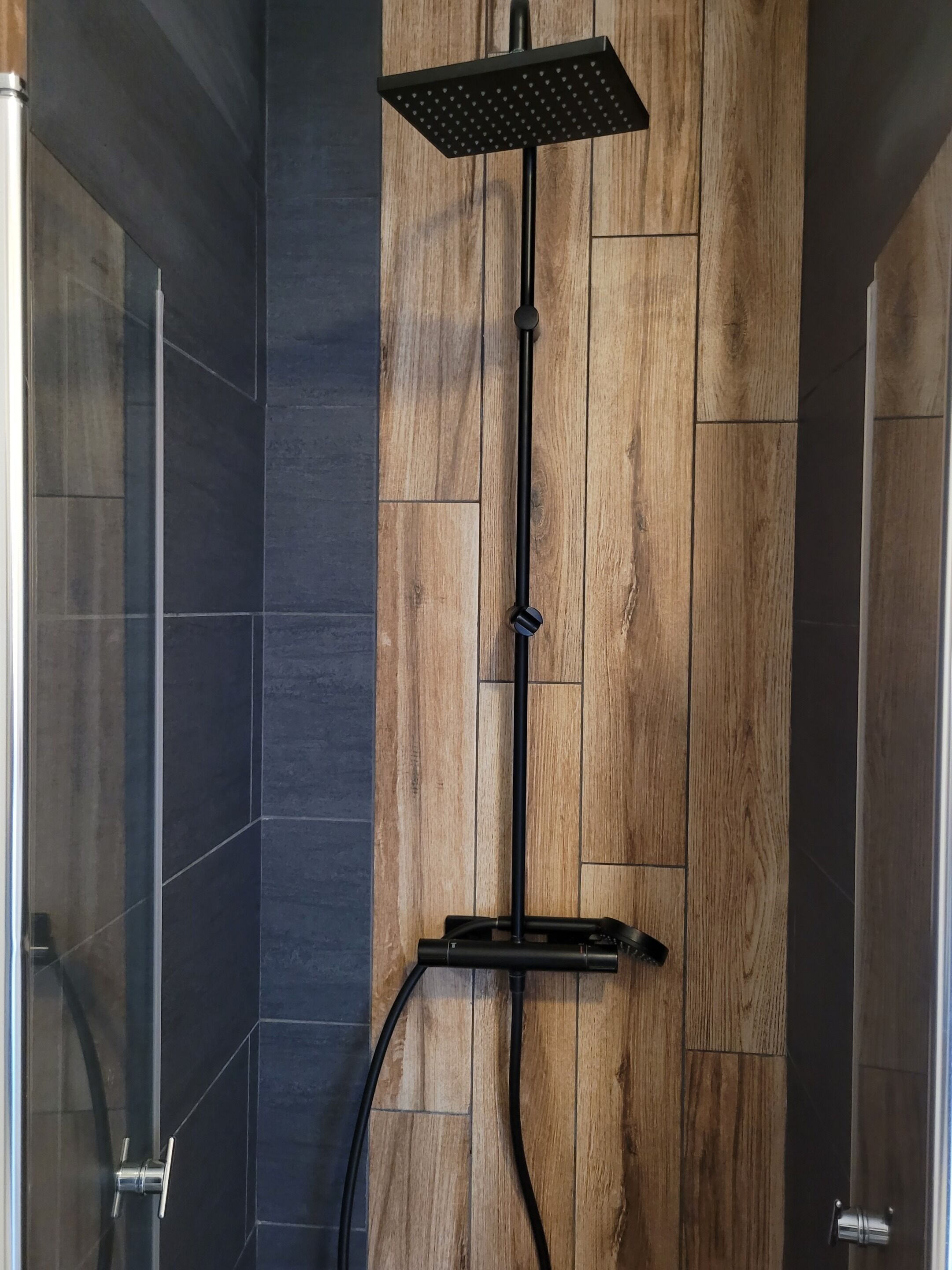 A shower with a wooden panel wall, a black showerhead, and a glass door.
