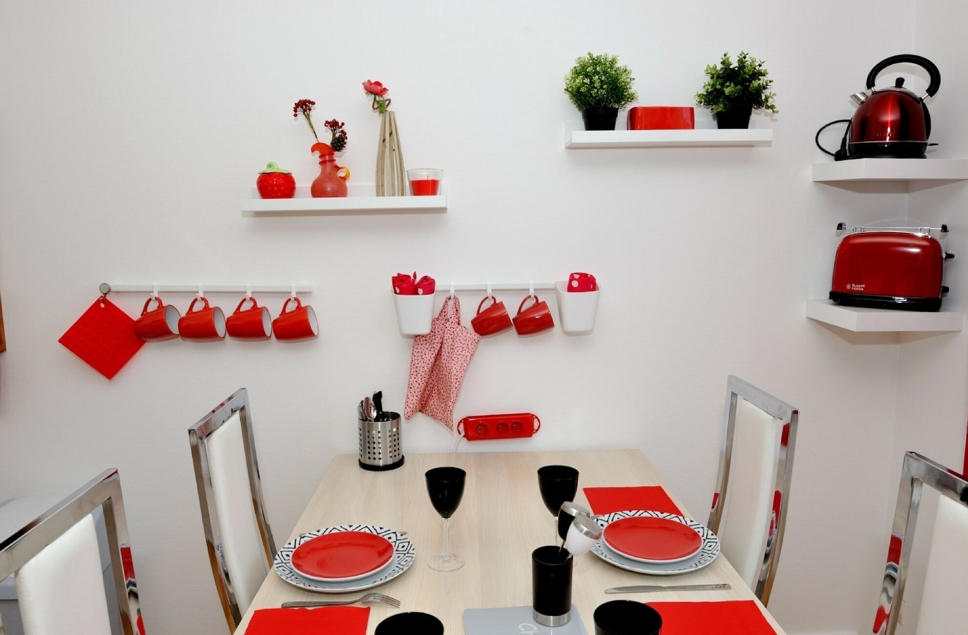 A dining area with a wooden table set for two, red and white plates, and a red and white napkin. There are red mugs on a wall-mounted rack, a red kettle, and a toaster on a shelf.