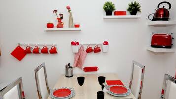 A dining area with a wooden table set for two, red and white plates, and a red and white napkin. There are red mugs on a wall-mounted rack, a red kettle, and a toaster on a shelf.