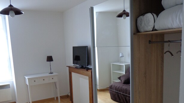 A room with a wooden floor, a white desk with a computer, a bed with a brown blanket, and a wardrobe with hanging clothes.