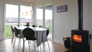 Un coin repas avec une table ronde, des chaises et une vue sur un pont, visible à travers de grandes fenêtres. Un poêle à bois noir est visible sur la droite.