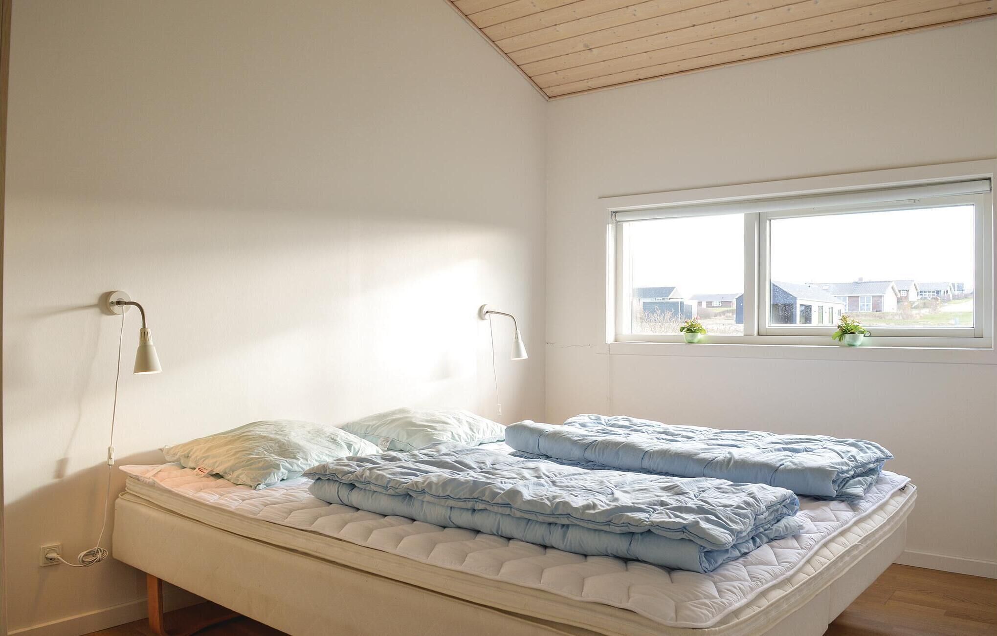 A bedroom with a bed, two wall-mounted lamps, a window with a view of houses, and a wooden ceiling.