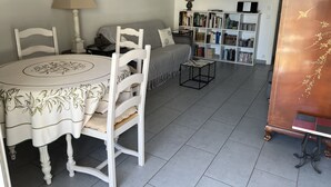 A dining area with a round table and chairs, a bookshelf with books, a bed with a grey blanket, and a decorative cabinet with a bird design.