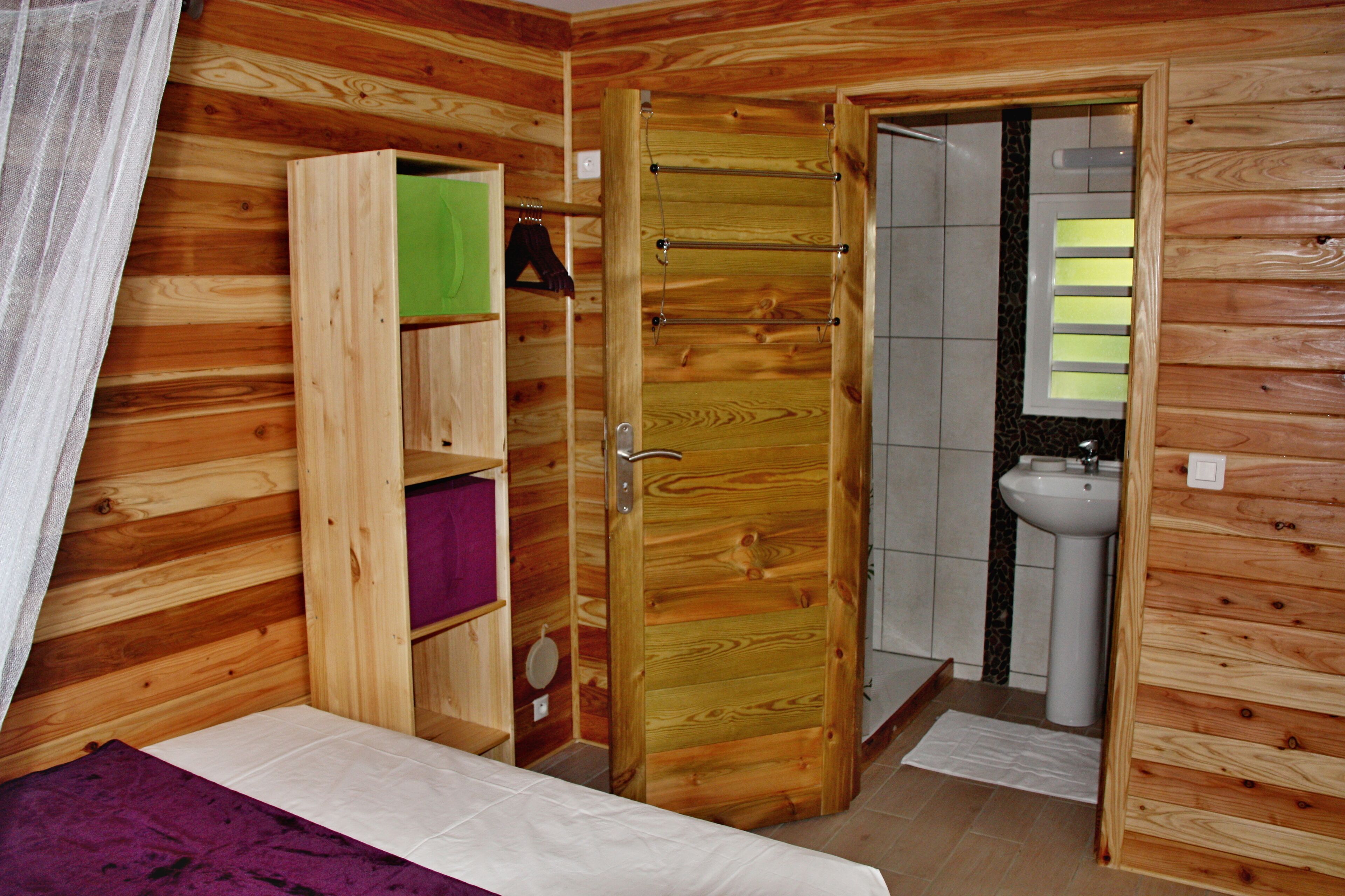 Una habitación de madera con una cama, un armario, un baño con lavamanos y una puerta que da a otra habitación.