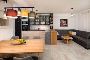 A modern kitchen with dark cabinets, a dining area with a wooden table and chairs, and a living room with a dark sofa and a coffee table.