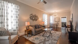 A living room with a sofa, armchair, coffee table, and patterned rug. There is a ceiling fan, a dining area with chairs, and a television on a stand. - Front Porch, Sunny Coffee Nook, Gorgeous Bathroom, One block to CSU & Old Town! (Fort Collins)