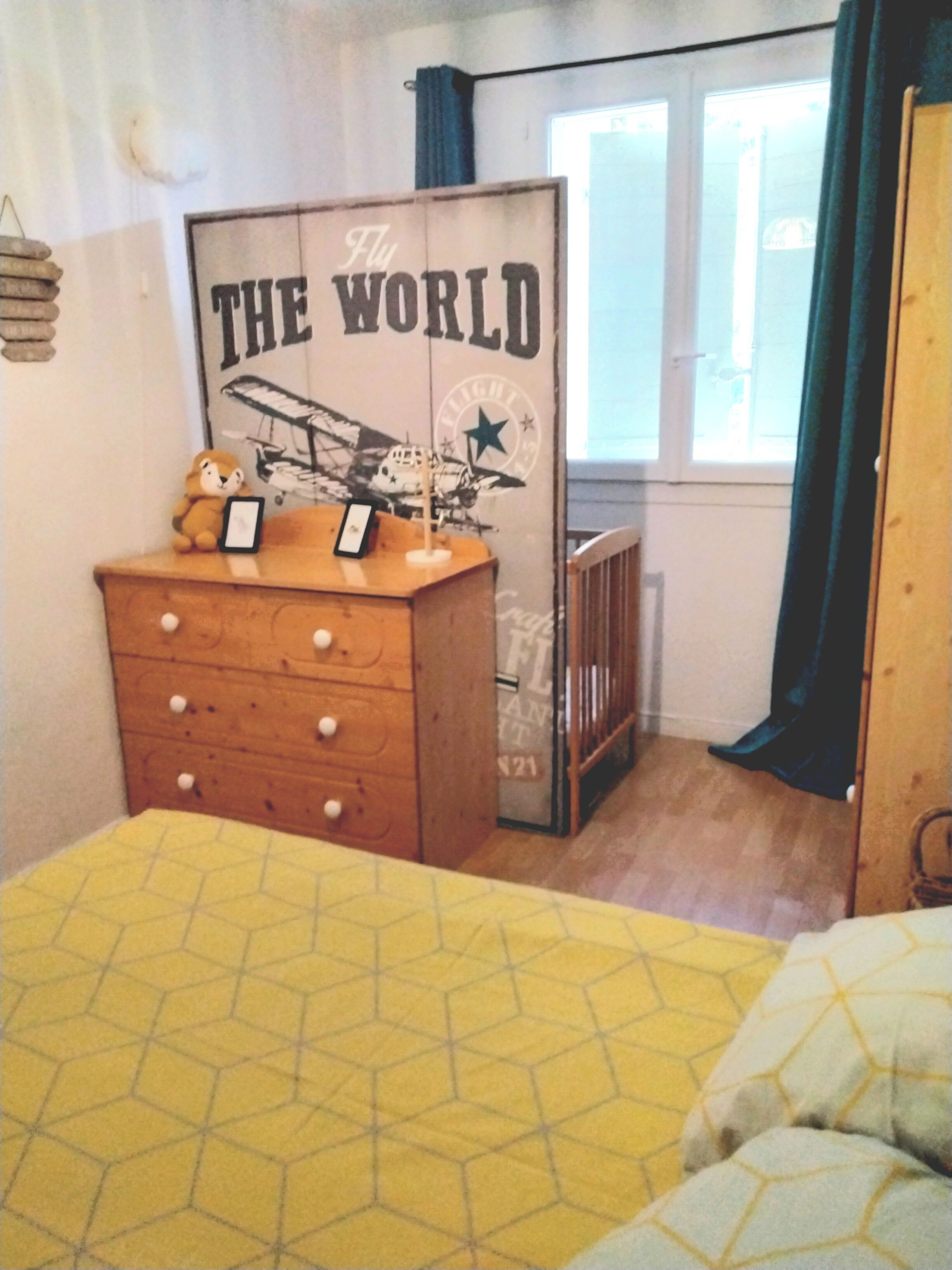 A bedroom with a wooden dresser, a bed with a yellow patterned bedspread, a crib, and a wall with a poster titled "THE WORLD".