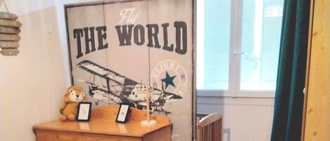 A bedroom with a wooden dresser, a bed with a yellow patterned bedspread, a crib, and a wall with a poster titled "THE WORLD".