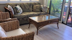 Uma sala de estar com sofá, duas cadeiras de vime, uma mesa de centro de vidro e uma porta de correr que dá para uma varanda.