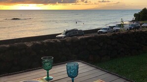 Mesa posta para dois, com pratos e copos, com vista para uma praia, carros estacionados e um pôr do sol.