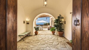 Un pasillo con suelo de piedra y un arco que da a un área exterior con sombrillas, plantas en macetas y vista al mar.