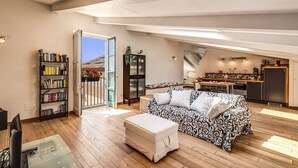 Un amplio espacio de estar con sofá, reposapiés y una estantería. Se ve una cocina al fondo.