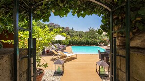Una terraza de madera que conduce a una piscina con sillas de descanso y sombrillas, rodeada de vegetación y un muro de piedra.