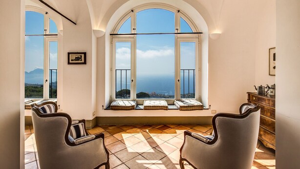 Una habitación con ventanas de arco que dan a vistas costeras, un armario de madera y dos sillones tapizados.