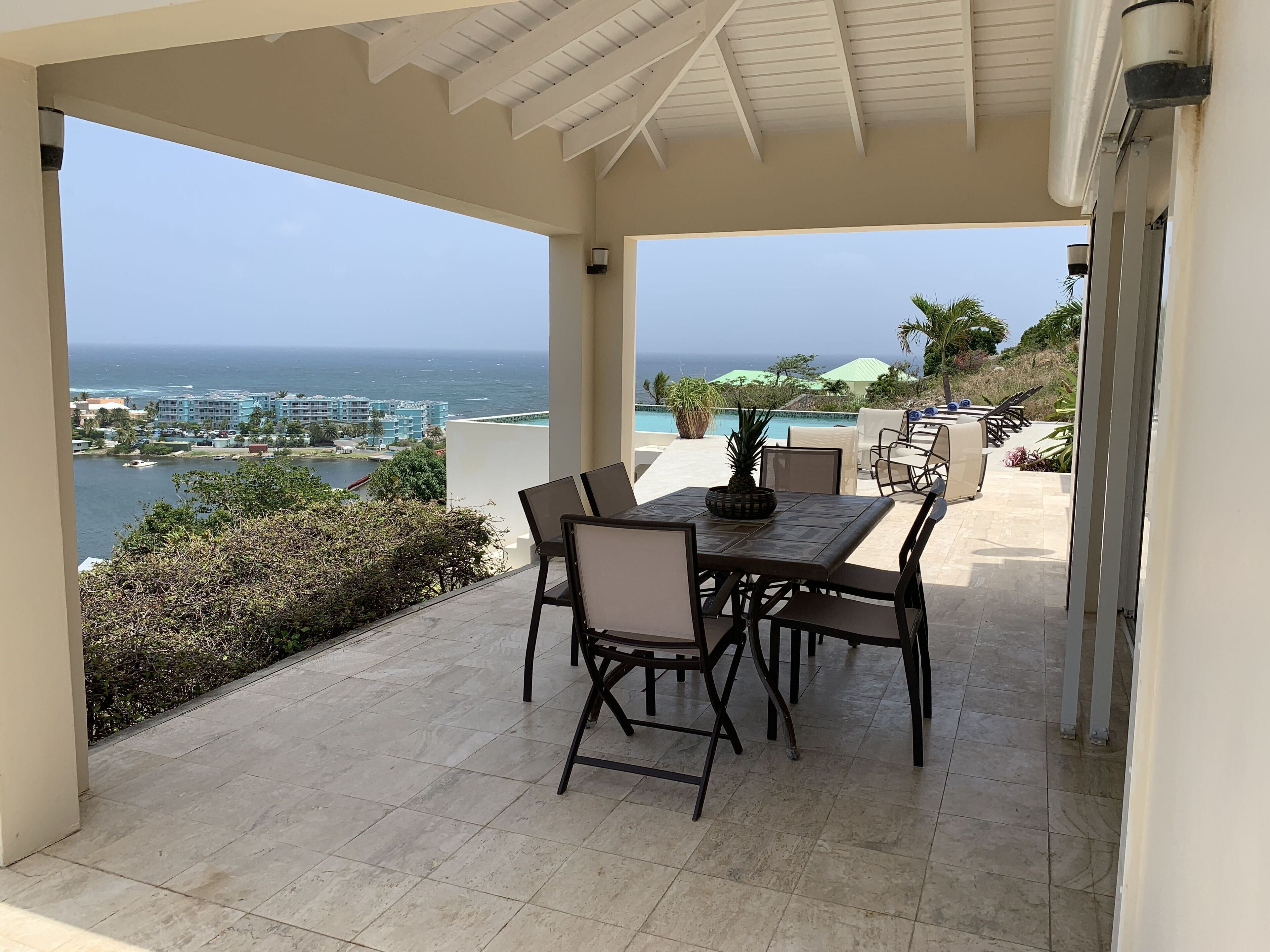 A covered outdoor patio with a table and chairs, overlooking a pool and ocean view.