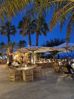 An outdoor dining area with wicker furniture and umbrellas, surrounded by palm trees.