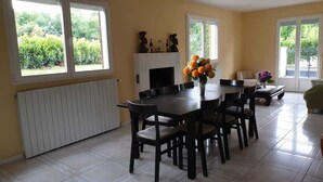 Un comedor con mesa y sillas, chimenea y ventanas amplias con vistas a la vegetación.