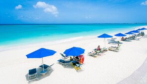 Una playa con arena blanca y agua turquesa cristalina. Se observan sombrillas azules y sillones de playa dispuestos a lo largo de la orilla.