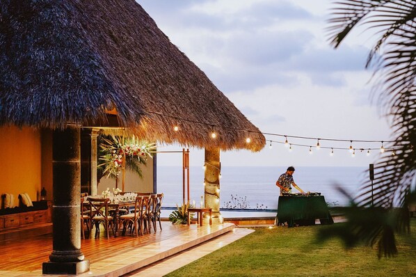 A thatched-roof outdoor dining area with a view of the ocean, a bartender at a bar, and string lights.