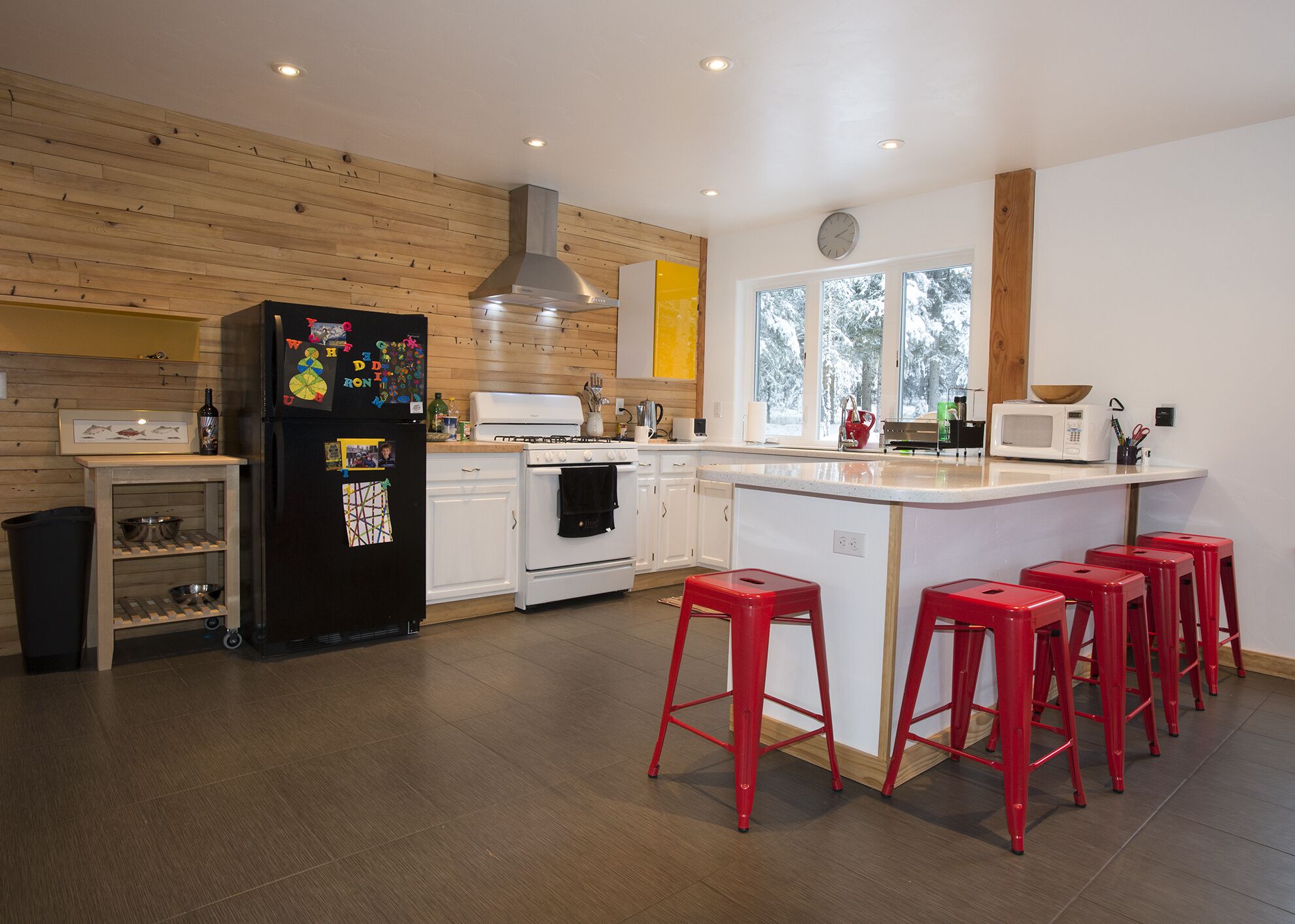 Una cocina moderna con paneles de madera, un refrigerador negro, armarios blancos, una estufa y taburetes de bar rojos.
