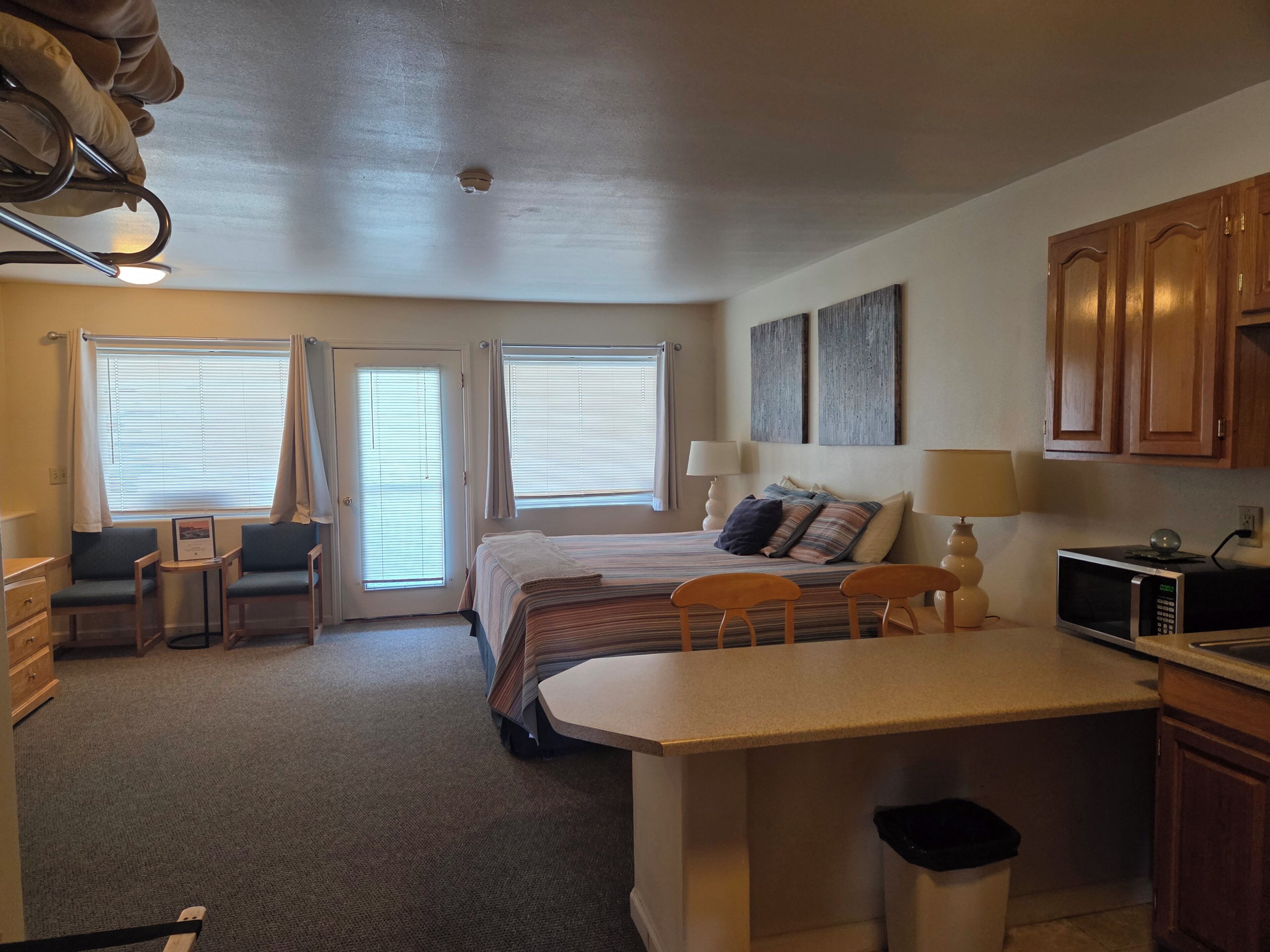 A compact hotel room with a bed, bedside tables, a small desk, a microwave, and a trash bin.