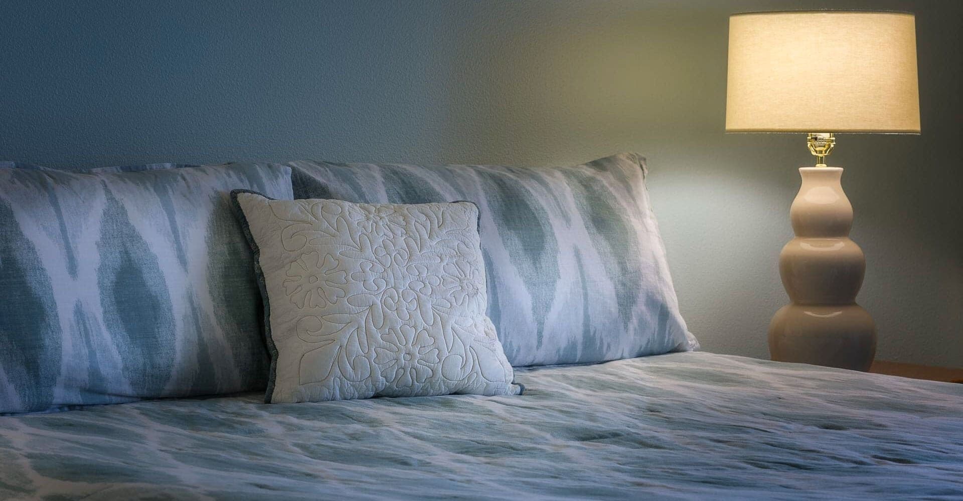 A bed with patterned bedding and two pillows, a bedside lamp, and a plain wall.