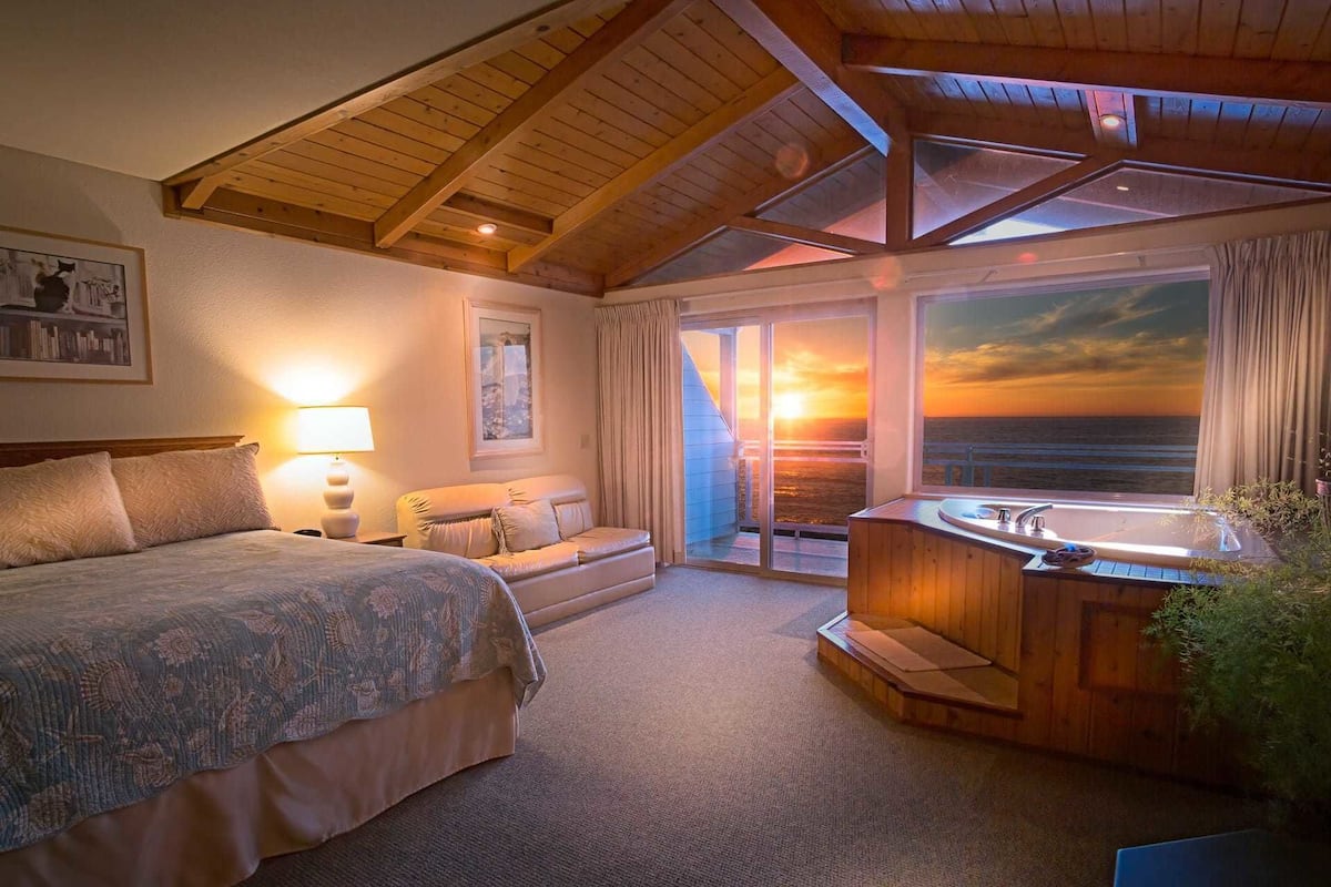 Ein Schlafzimmer mit einem großen Bett, einem Nachttisch, einem Schreibtisch und einem Fenster mit Blick auf einen Sonnenuntergang.