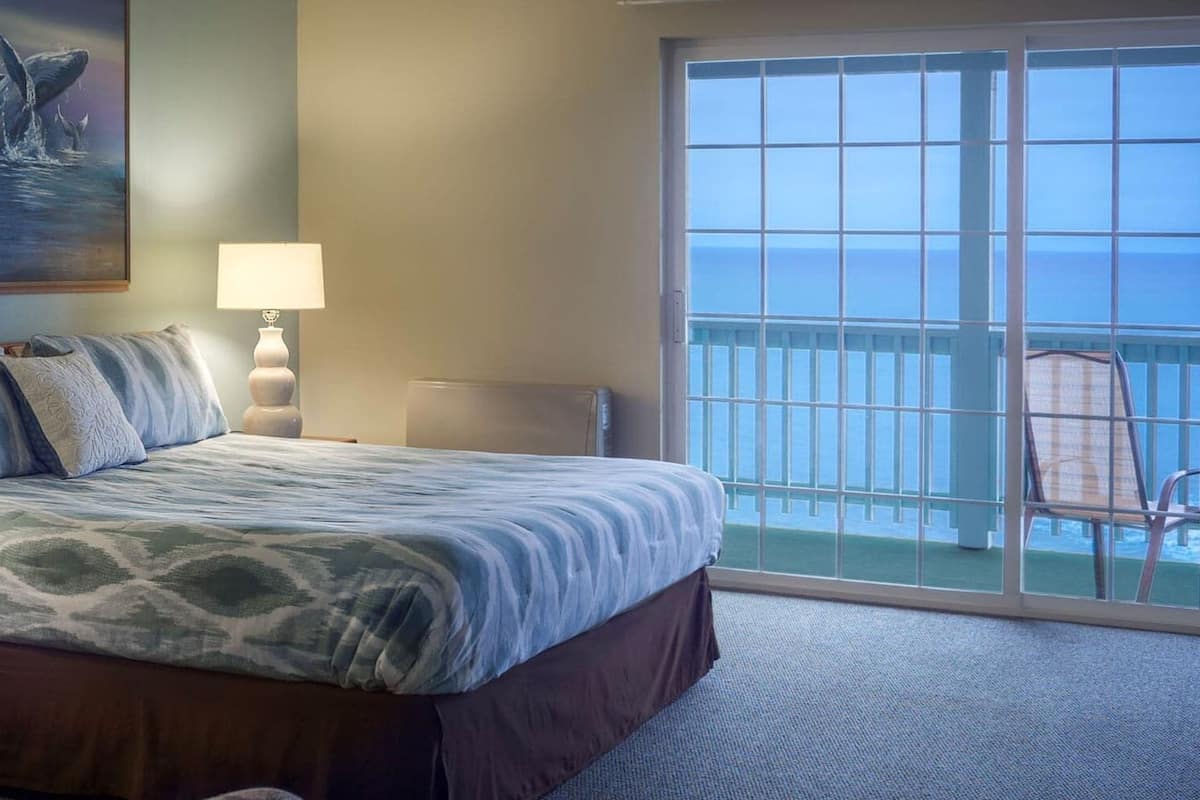 Ein Hotelzimmer mit einem Bett, einem Nachttisch, einem Walgemälde, einem Balkon mit einem Sessel und Blick auf den Ozean.