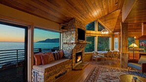 A cozy living room with a stone fireplace, large windows overlooking a body of water, and a dining area with wooden furniture. - Inn Of The Lost Coast (Shelter Cove)